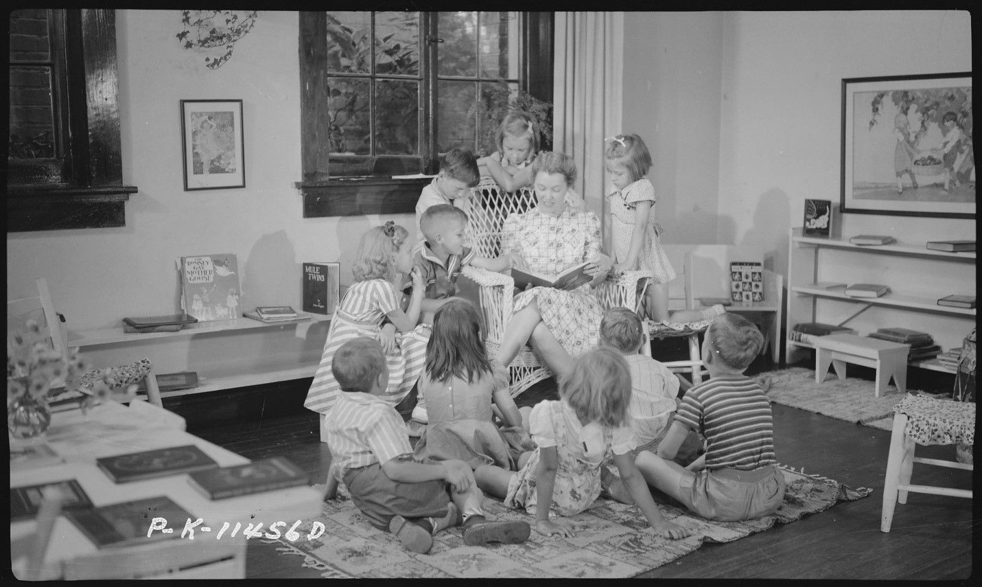 An archival photograph of a lady reading from a book, surrounded by many young children.